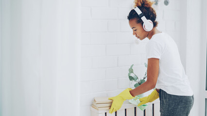 Cheerful maid African American girl is doing housework dusting using cloth and wearing protective...