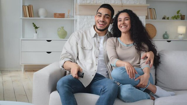 Hispanic Indian Latina multiracial married couple husband and wife man and woman rest at home on couch weekend switch channel with remote control choose TV program laugh smile watching movie film
