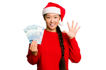Young asian woman celebrating Christmas holding banknotes isolated smiling cheerful showing number five with fingers.
