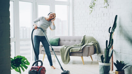 Beautiful young woman is hoovering the floor at home using modern vacuum cleaner and listening to music with headphones, dancing and singing. Housework and technology concept.