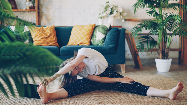 Flexible young blonde is doing stretching exercises at home sitting on floor in modern apartment. Girl is bending head to knee holding toes and stretching back and legs.