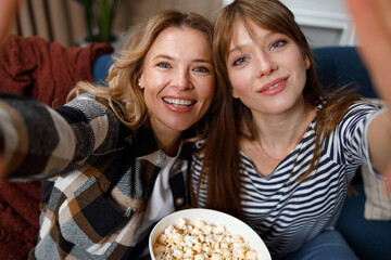 Two woman of different ages take a selfie sitting on the couch with a bucket of popcorn watching a...