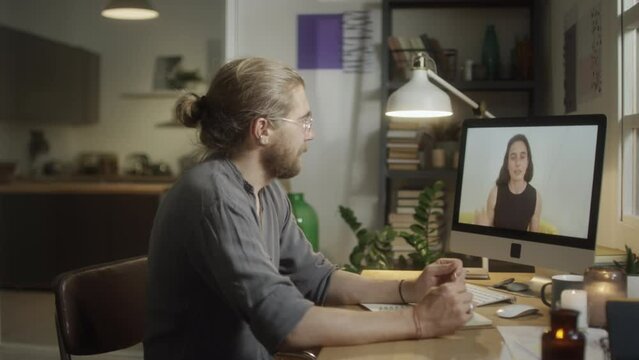Medium shot of man listening to female tutor via video call on computer while taking online class in evening at home