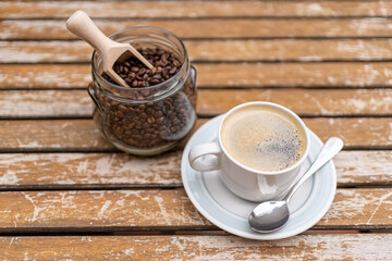 Coffee cup with glass jar filled with coffee beans on a wooden garden table.