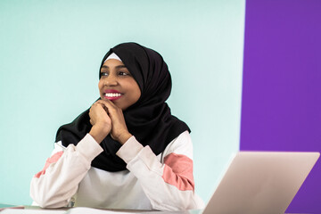 Afro woman wearing a hijab thoughtfully sitting in her home office and uses a laptop and smartphone 