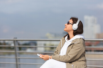 Attractive woman with headphones and mobile in hand sits on the city embankment and enjoying music...