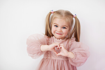 Little girl in pink dress with ponytails shows heart sign with her hands isolated on white background