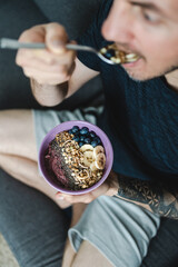 Man sitting on the couch while eating muesli and smoothie bowl with fruit and nuts for breakfast....