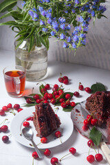 Chocolate cake with sweet cherry, cup of tea and bouquet of blue flowers on white background..