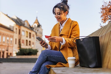 Young adult woman sitting on a bench does some online shopping. Woman in a yellow coat holding a...