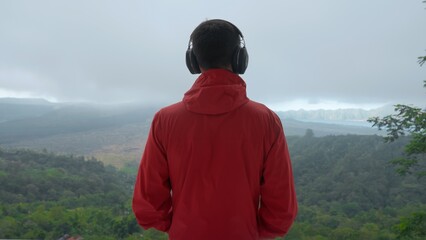 Adult man listening to music through large headphones in mountains on a stormy day looking at valley and misty volcano. Music-lover on a hiking trip wearing red jacket enjoying mount view. Soft focus.
