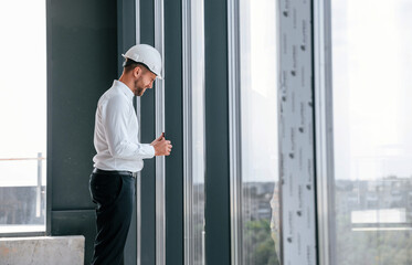 Standing near panoramic windows. Man in white shirt is working indoors at construction site. Managing the project