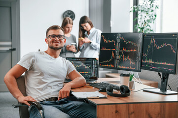Stock broker is sitting by computer and working with exchange technology. Women is standing and...