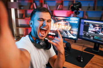 Making selfie, celebrating victory. Male game streamer in casual clothes is indoors with pc