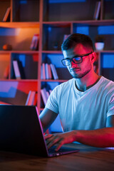 Focused by work. Man in glasses and white shirt is sitting by the laptop in dark room with neon...
