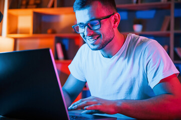 Positive emotions. Man in glasses and white shirt is sitting by the laptop in dark room with neon...