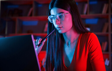 Holding stylus. Beautiful woman in glasses and red wear is sitting by the laptop in dark room with...