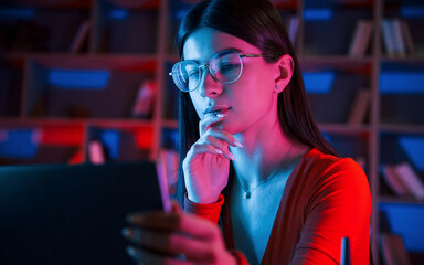 Beautiful woman in glasses and red wear is sitting by the laptop in dark room with neon lighting - obrazy, fototapety, plakaty