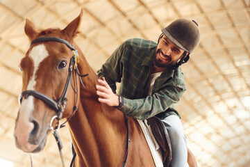 Sitting in the saddle. Young man with a horse is in the hangar