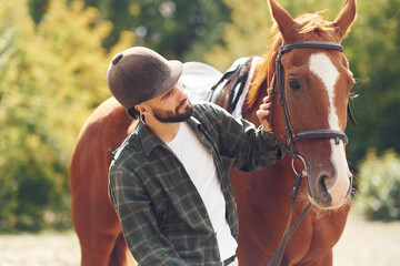 Fototapeta premium In protective hat. Young man with a horse is outdoors