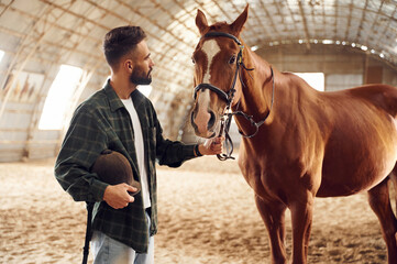 Standing and holding animal. Young man with a horse is in the hangar