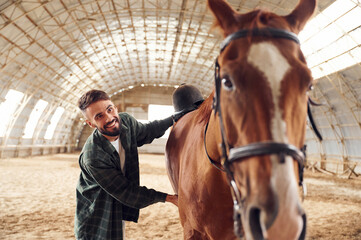 Smiling and taking care of animal. Young man with a horse is in the hangar - obrazy, fototapety, plakaty