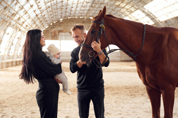 Lovely couple is standing with indoors in the hangar with little baby and showing him horse
