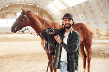 Obraz premium Holding the saddle in hands. Young man with a horse is in the hangar