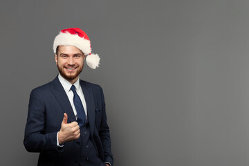 Portrait of cute business guy successful businessman wearing suit and Christmas hat showing thumb up against grey studio wall background.