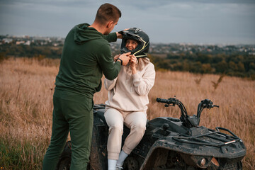 Man is helping his girlfriend to wear a helmet. Lovely young couple is with quad bike outdoors on the field © standret