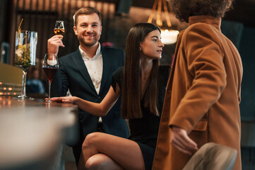 Woman and man sitting in the bar with drinks and welcoming their friend