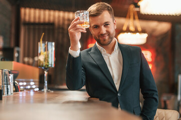 Sitting by the table. An elegant man in black suit is in the bar, tasting the alcohol drink