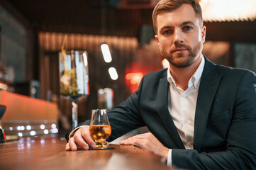Sitting by the table. An elegant man in black suit is in the bar, tasting the alcohol drink