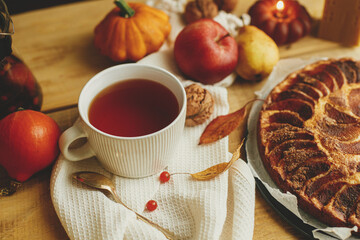 Atmospheric autumn still life. Warm tea in stylish cup on rustic wooden table with freshly baked apple pie with cinnamon, candle, autumn leaves and nuts. Hygge home, cozy moment