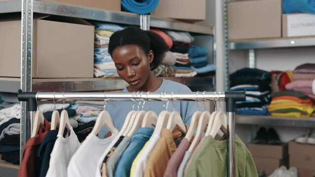 African american female volunteer goes over hangers with clothes for recycle while working at the second hand or charity organization. Reuse, second-hand, conscious consumer, sustainability concept