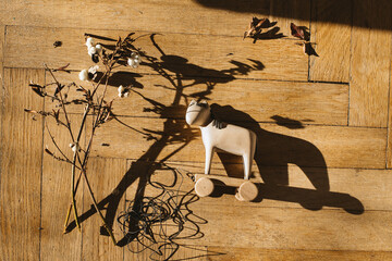 A lonely wooden toy horse lies on the wooden floor amidst dry branches. In the sunlight, it reflects the end of childhood and the fleeting comfort, embodying the sense of abandoned play and solitude