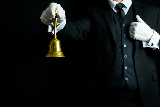 Portrait Of Butler Or Waiter In Dark Suit And White Gloves Holding Gold Bell On Black Background. Service Industry And Professional Hospitality.