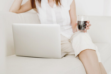 Fototapeta na wymiar Cropped image of businesswoman holding cup of coffee and working with laptop while sitting on sofa in living room.