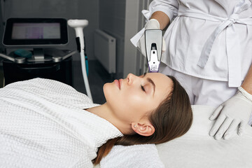 Pretty woman enjoying radiofrequency lifting procedure for her face and forehead skin rejuvenation in aesthetic cosmetology. RF lifting - 542738543