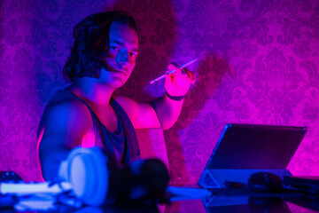 Handsome young guy is sitting with a digital pencil in front of tablet at table, the room is illuminated by a muted violet-blue light. Creative process of artist who has caught inspiration.