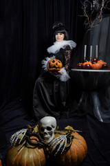 A beautiful brunette in a vintage black dress, a white frill and a vintage hat surrounded by pumpkins and skulls. Happy halloween holiday concept. - obrazy, fototapety, plakaty