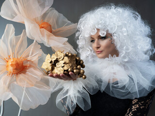 A beautiful girl in a vintage black dress, a white frill and a lush curly wig poses in a studio with huge flowers. Carnival costume. - obrazy, fototapety, plakaty