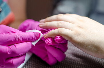 Female manicurist in pink gloves works with young hands and nails under a bright lamp. Process of wiping the nail with a lint-free cloth from dust