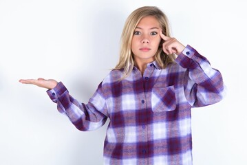 Obraz premium beautiful caucasian teen girl wearing plaid purple shirt over white wall confused and annoyed with open palm showing copy space and pointing finger to forehead. Think about it.