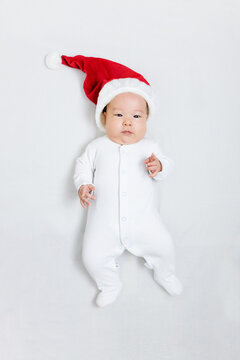 Asian Baby 5 Months Old In A Santa Hat And White Suit Lies On A White Background, Pointing Gesture