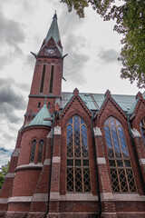 Obraz premium Finland, Kotka - July 18, 2022: Kotka-Kymin Parish Church or Seurakuntayhtymä. North red brick facade with clock tower against gray cloudscape. Gothic-revival windows and green roof