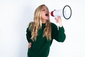 Funny beautiful caucasian teen girl wearing green knitted sweater over white wall People sincere emotions lifestyle concept. Mock up copy space. Screaming in megaphone.