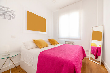 Bedroom with double bed with yellow velvet cushions, glass bedside tables and pink bedspread and white framed mirror