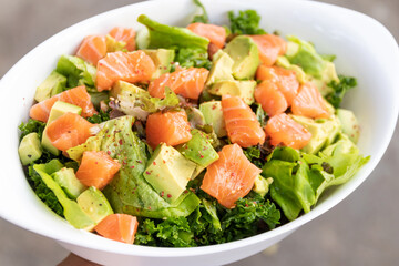 Salmon salad with green leaves and avocado on oval white bowl.