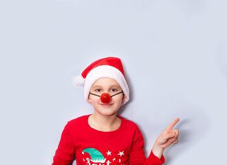 Cute caucasian kid boy in red Santa's hat and red nose points with his finger on empty copy space for design or text. Banner. Christmas mockup template for card or certificate.